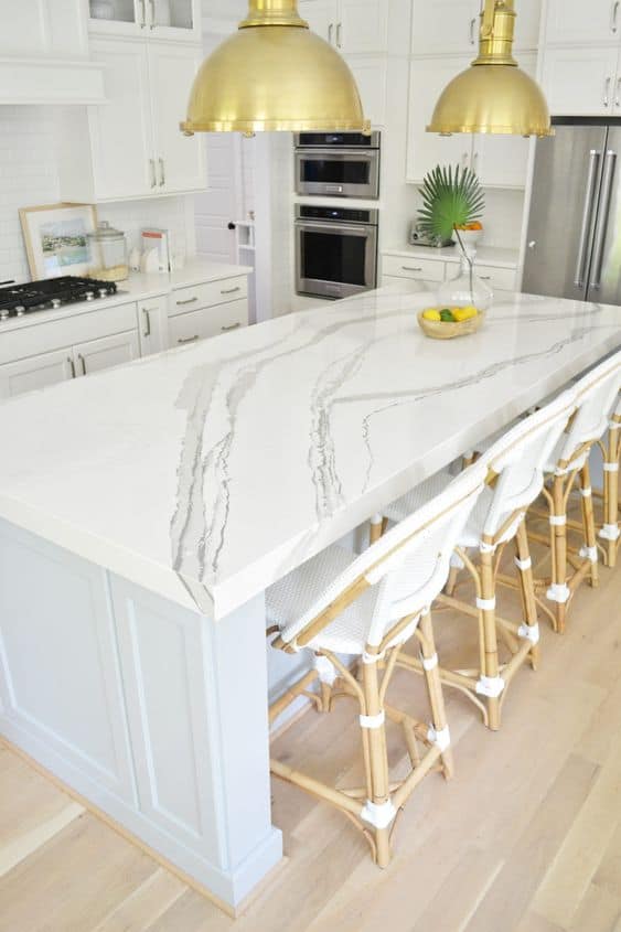 white and grey textured quartz countertop designs for kitchen with white and brown chairs, golden pendant lights, and wooden laminate flooring