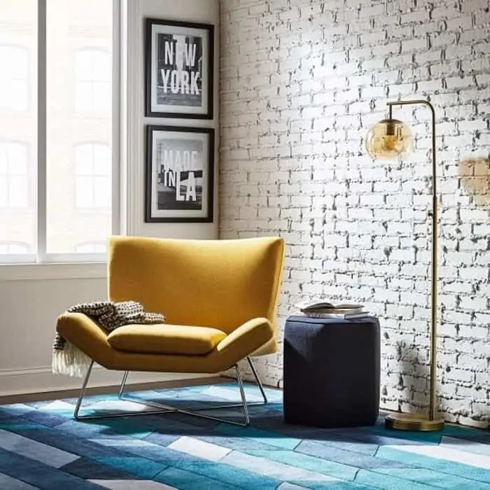 Yellow accent chair placed in a living room with small blue stand, a lamp, some wall paintings and a geometric patterned rug underneath.