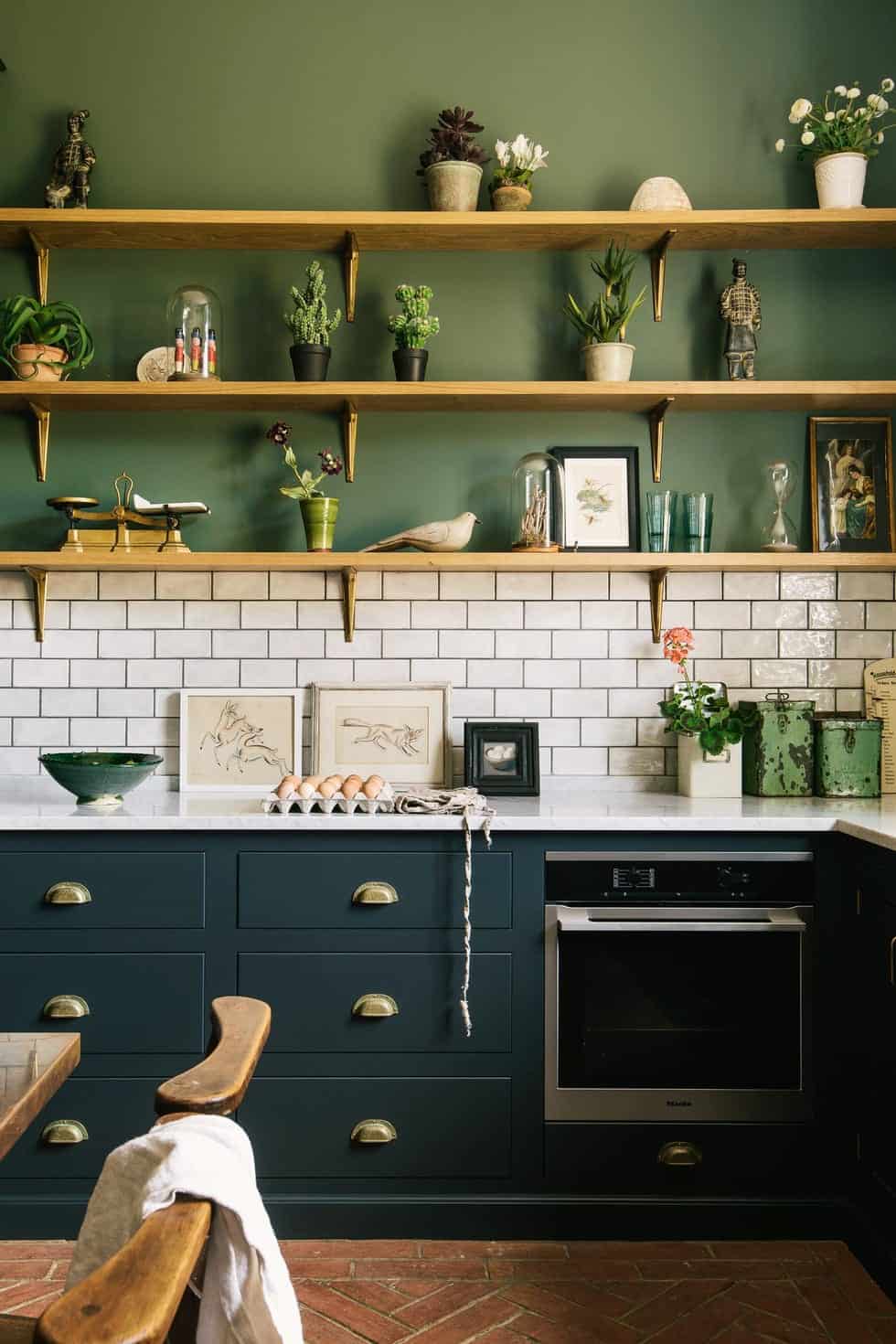 kitchen with green walls and white metro tiles and wooden shelves with plants and artwork