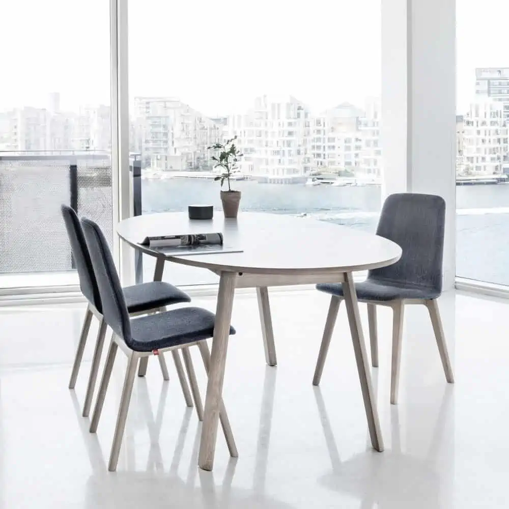 A white top dining table with light wooden frames, 3 chairs with dark gray seating in a room with huge windows