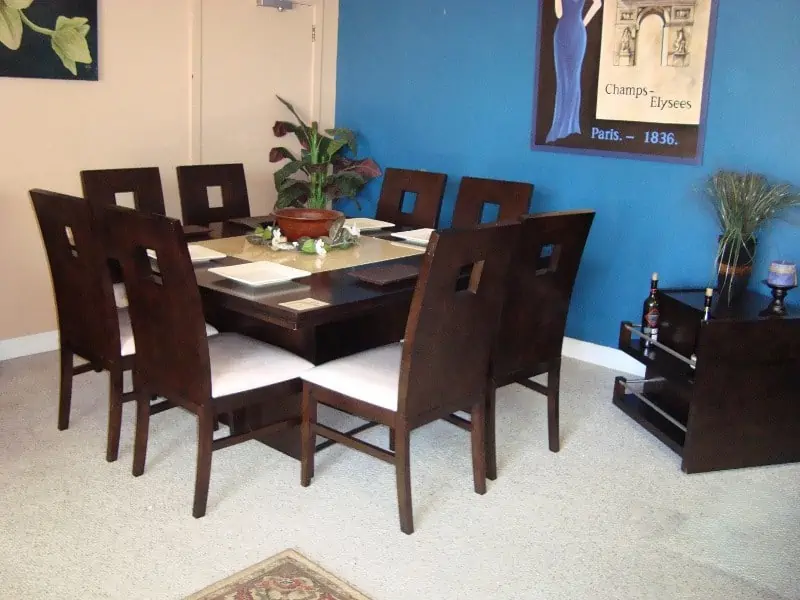 Square 8 seater dining table, wooden chairs with white cushions, dark brown, in a room with blue and light orange walls