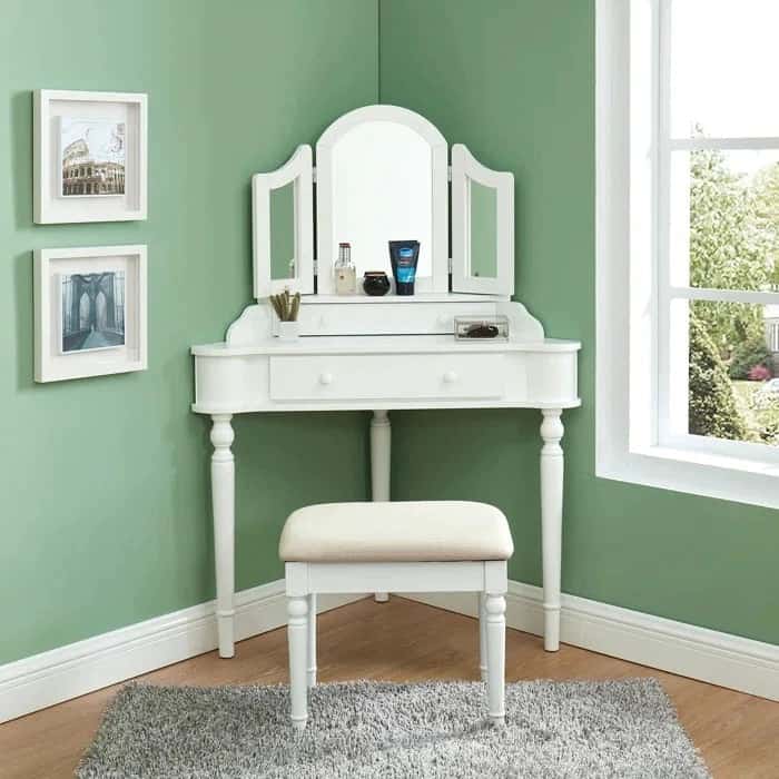 A pretty, white-colored, wooden dresser with small 3-fold mirrors, minimum storage space, and a matching stool, in a green room.