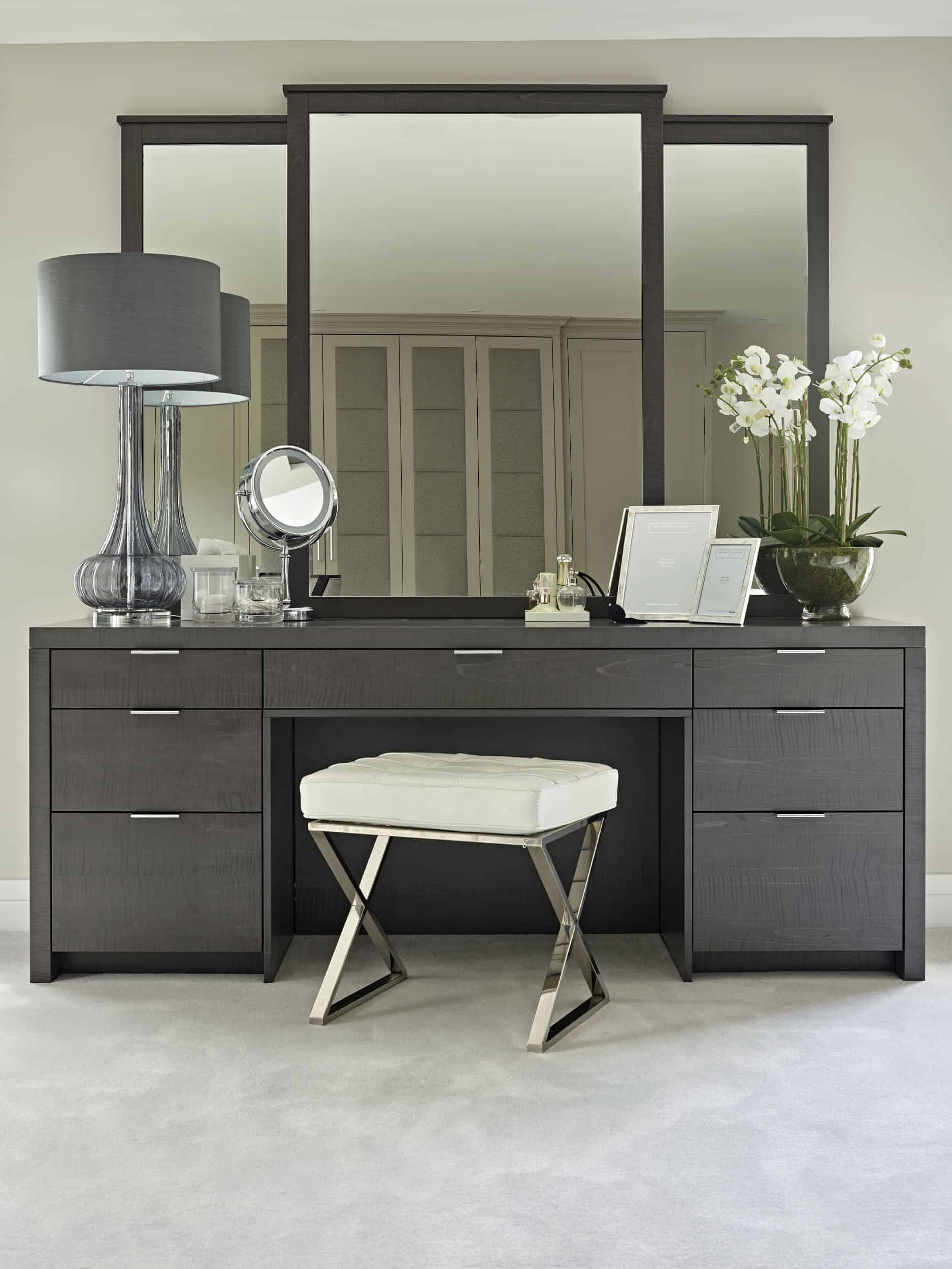 A sturdy black-colored dresser with a huge 3-fold mirror, lots of storage space, and a matching stool, in a white room.