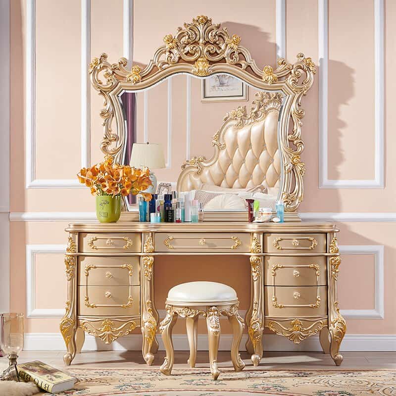 An exquisite royal golden-colored dressing table design with a huge mirror, lots of storage drawers, and a matching stool, in a peach-colored room.