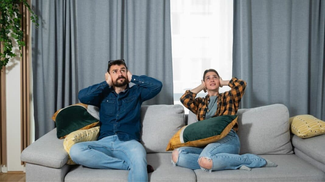 a couple shutting their ears to avoid noise, sitting on a grey sofa