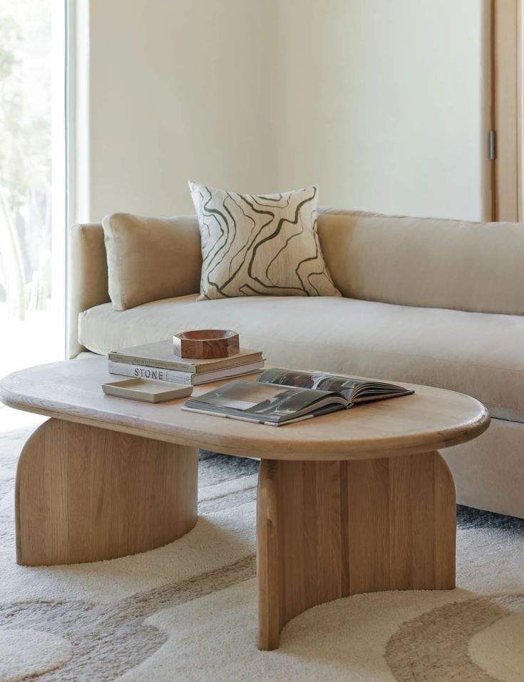 a wooden oval shaped centre piece, with a rustic element, beige-white sofa and cushions, beige carpet, with beige walls, adding a minimal vintage touch to your space, magazines, books