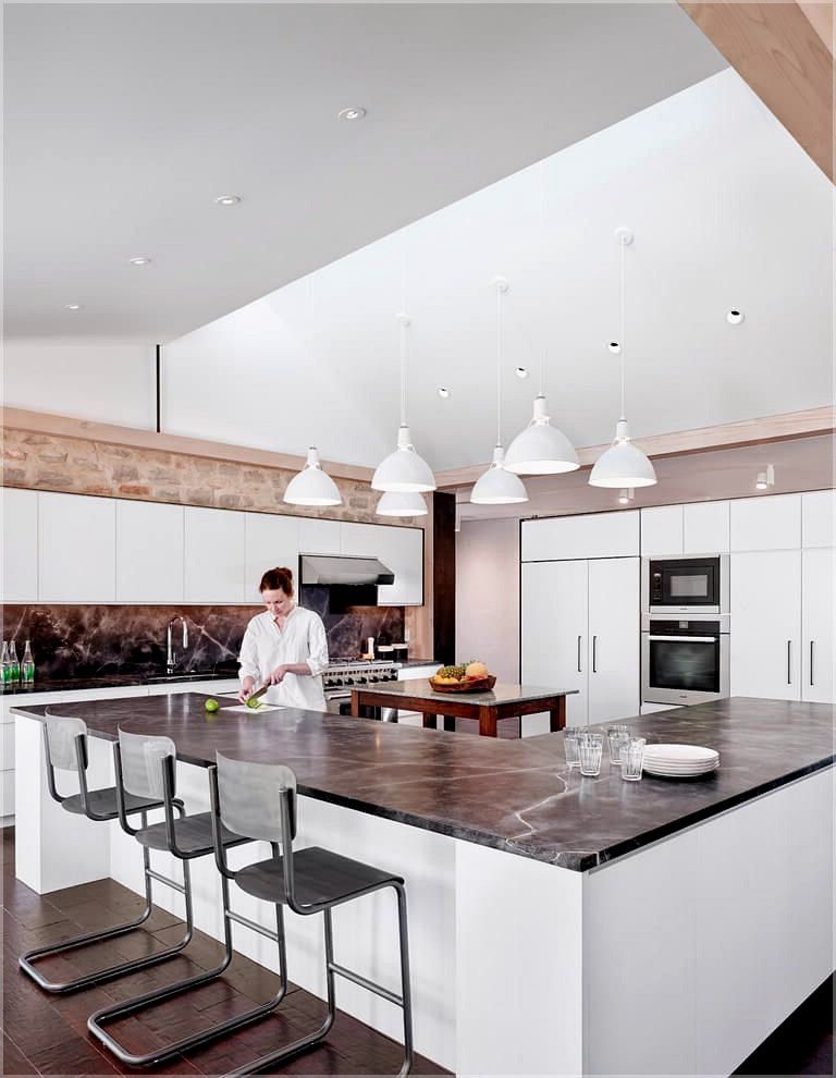 L-shaped Island kitchen design with seating, white pendant lights hanging above the countertop, marble countertop, wooden table in the centre, in-built oven in the cupboard, white island body