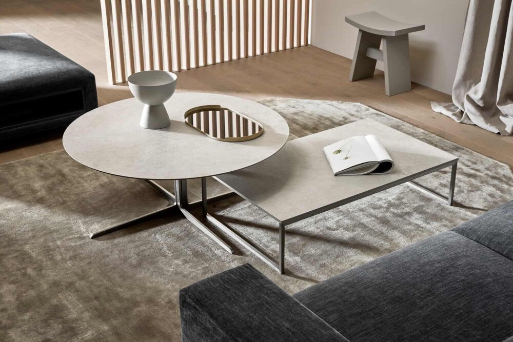 modern round centre piece in a living room with a beige tabletop, and a complementary rectangular centre piece which has a metal base, wooden flooring, grey sofa, beige walls, classic living room
