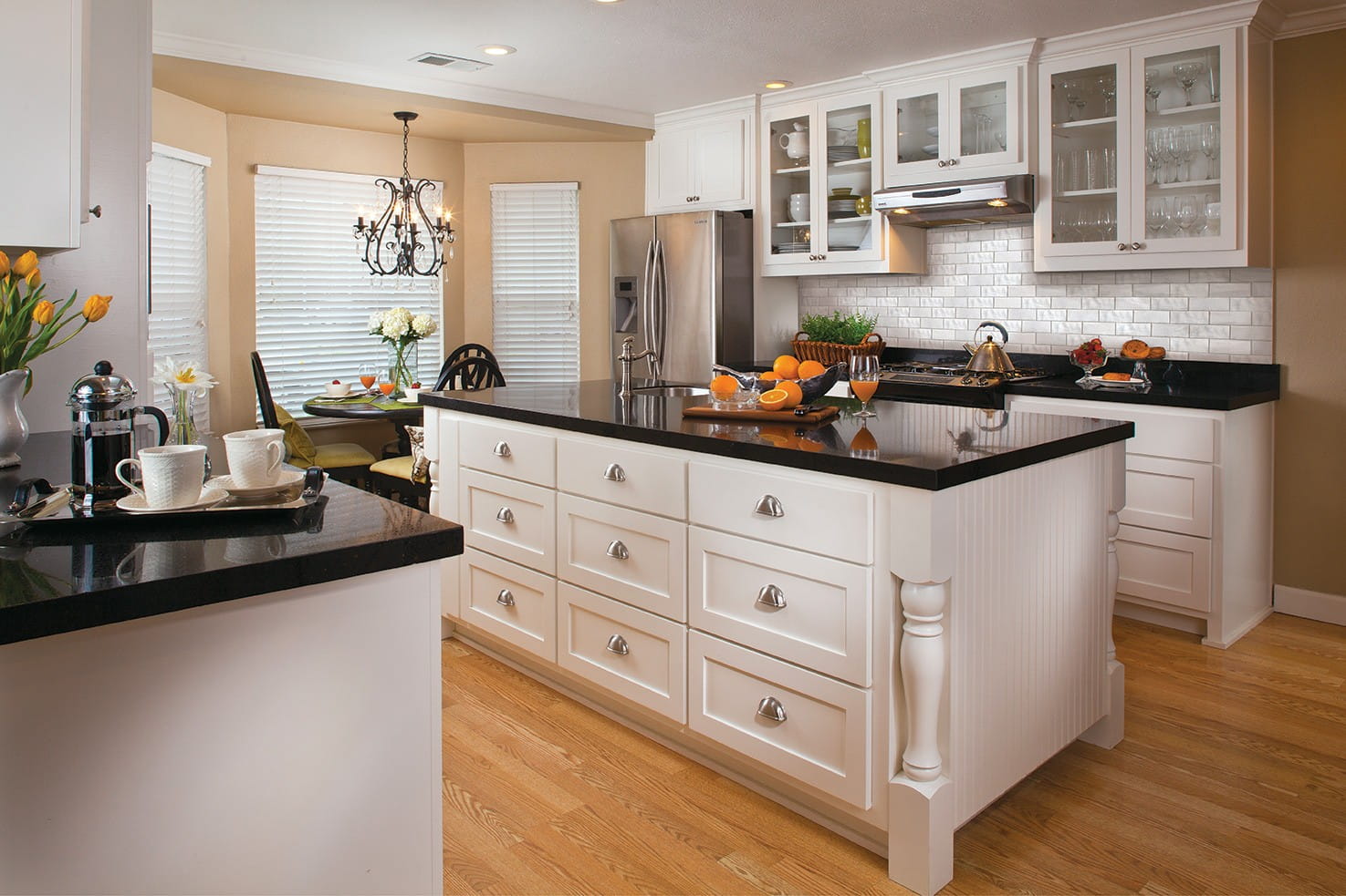 a beautiful galley kitchen with a centralised island, having cabinets and its countertop has a black marble finish, the cabinets are white, the island countertop has a built-in sink, it is a storage efficient kitchen with above cabinets too, seating arrangement with a table and chairs, chandelier installed above the seating area