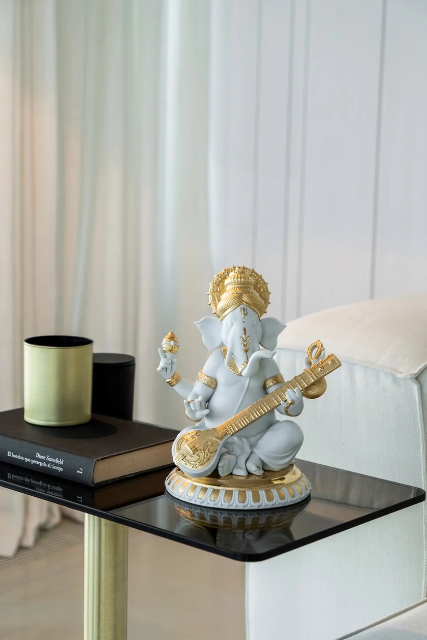 Ganapati statue placed on a glass table, matte white porcelain finish with touches of gold lustre decorating various parts of the piece, beautiful decor piece