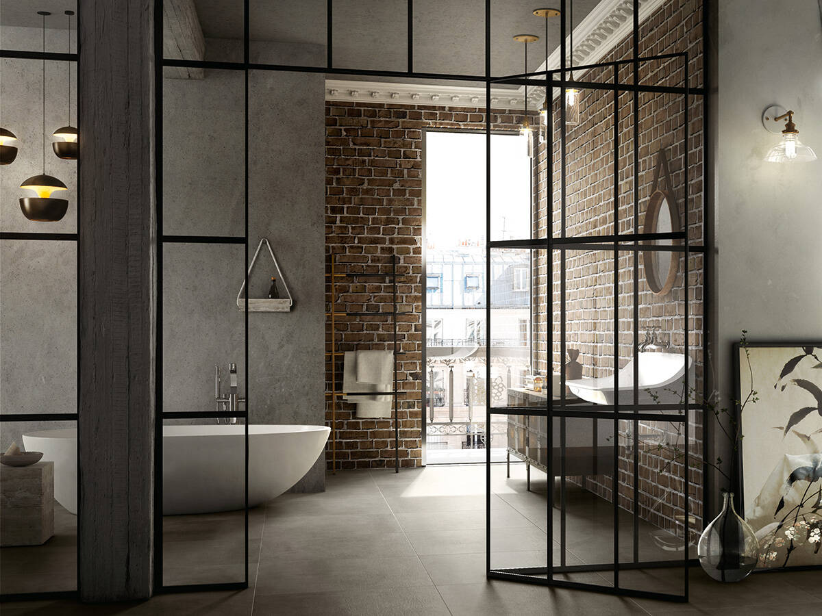 grey walls in a washroom with a tub, washbasin, mirror and reddish brown exposed brick wall