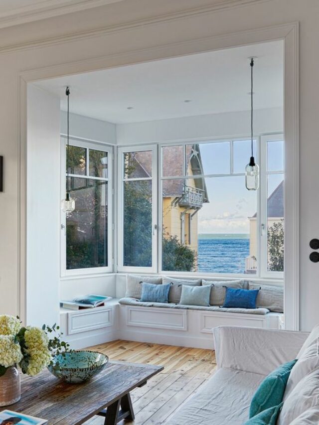 a-beautiful-coastal-living-room-with-a-bow-window-that-shows-off-amazing-sea-views-and-a-windowsill-bench-with-pillows