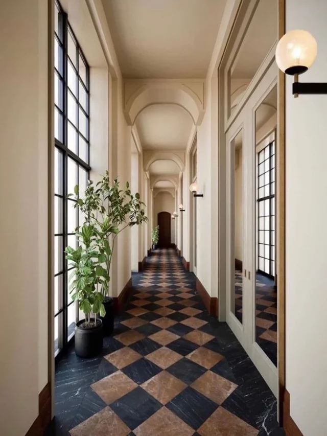 Hallway with beige walls, brown and black checkered floor, black pots on the side with indoor plants, wall lamp