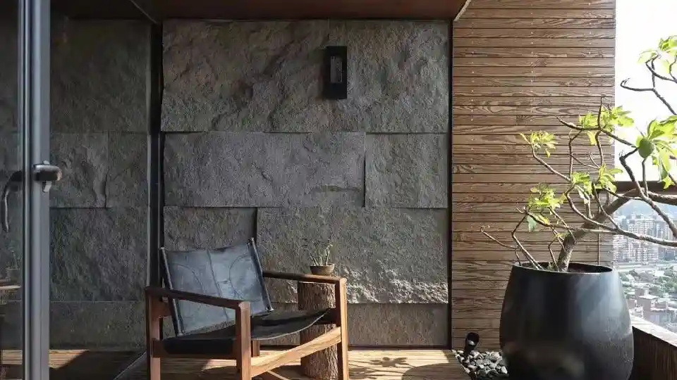 a grey exterior rock tiles textured wall in a balcony, a modern rocking chair and potted plants, and a wood log side table with a small potted plant on top