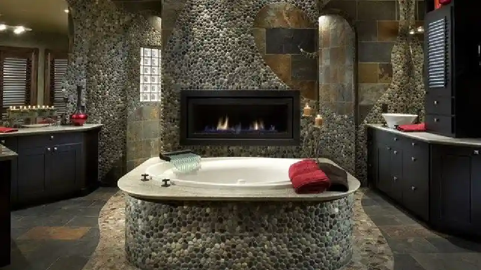 A traditional-looking bathroom design, an inset bathtub with a pebbled rock tile wall, fireplace and cabinets in the background