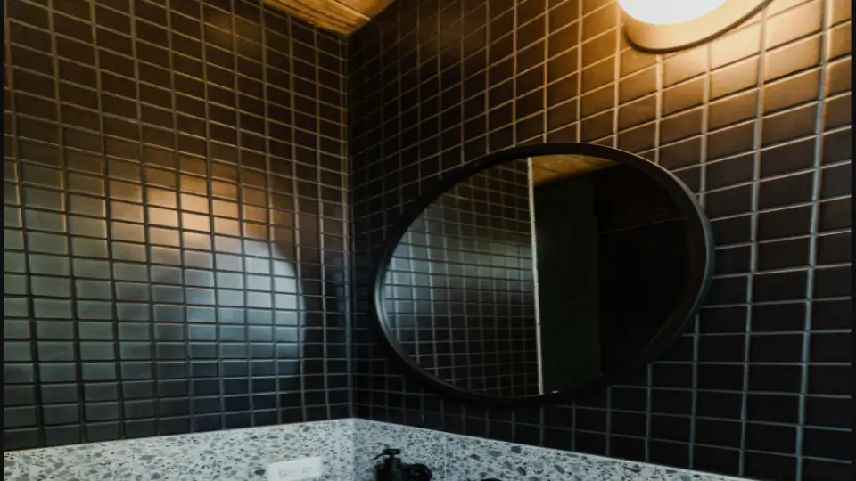 Small mosaic tiles in bathroom with yellow light on top and a circular mirror with black rim