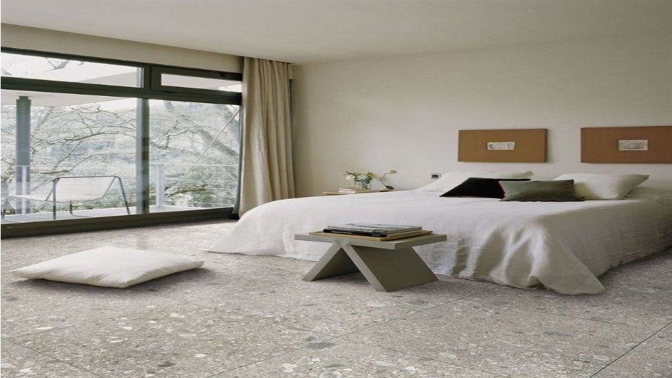 bedroom with windows, a bed with white cover, grey table on it are books, a pillow is on the floor, the view outside ia of a dried tree branches and it is snowing