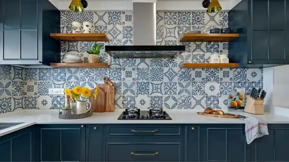 Blue decorative tiles for walls, dark blue cabinets, dark brown floating shelves with decor pices on them, silver electric chimney, white countertop with in-built gas stove, and other kitchen utilities like knife stand and chopping board