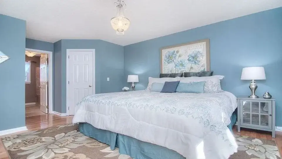 As per west facing house Vastu plan, the master bedroom has a bed covered with white blankets. There is a lit table lamp on the side table next to the bed. The walls are painted blue and a chandelier hangs at the centre of the ceiling.