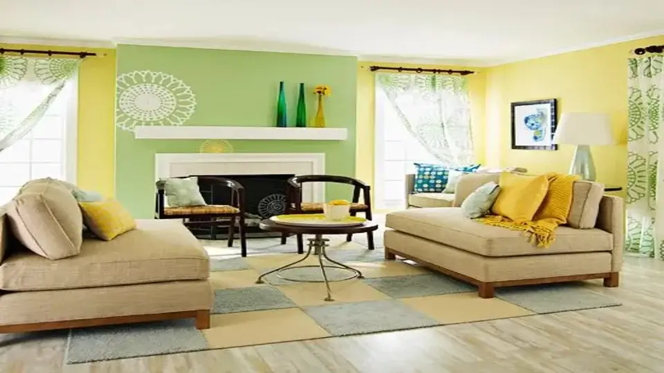 According to Vastu in west facing house, the living space featuring walls painted in pastel shades of yellow and green. There is a seating arrangement along with a centre table. The large windows allow natural light, partially covered with sheer screens. 