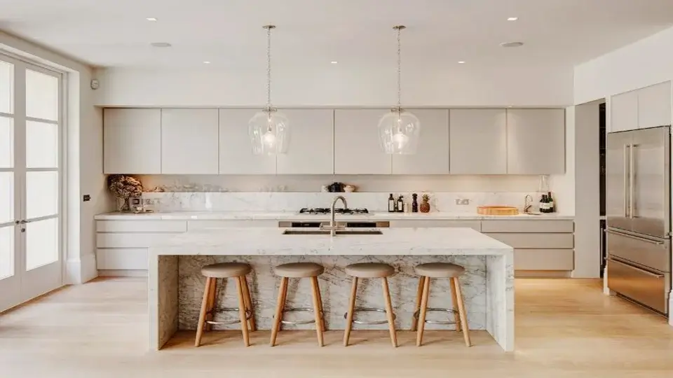 The kitchen is painted white with a counter in the centre. There are four bar stools and a refrigerator on the side. Pendant lights hand over the counter-top.