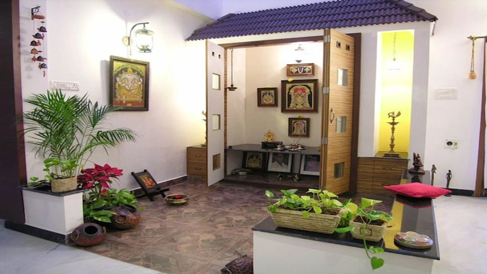 West facing house Vastu plan with Pooja room is adjacent to the living room and an idol placed within. Wooden doors cover the outer area. Spacious space in the front is covered with indoor pants.