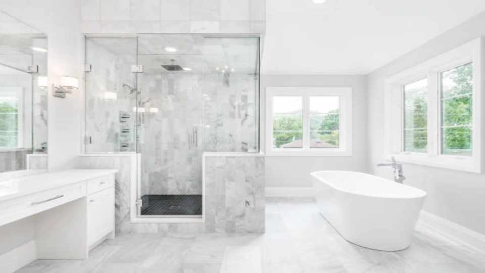 The bathroom features vanity parallel to the bathing tub. The shower area is enclosed with glass doors. The bathroom is predominantly white in color with marbles on the floor and on the wall.