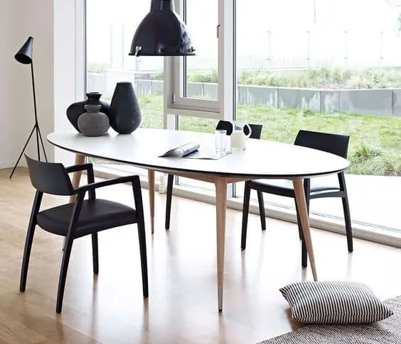 oval white table top with wooden legs, black chairs, in a room with large window, ceiling light and wooden floor