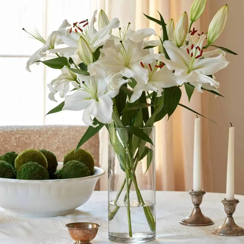 White lily flowers in a glass vase with green foilage, candles and a bowl of fruit