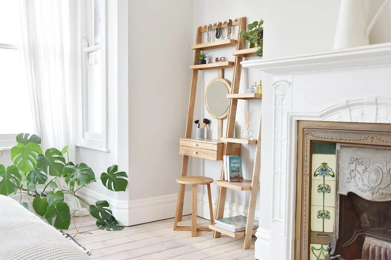 A wooden ladder-style dressing table design with a round mirror in a well-lit bedroom with plants and a fireplace.