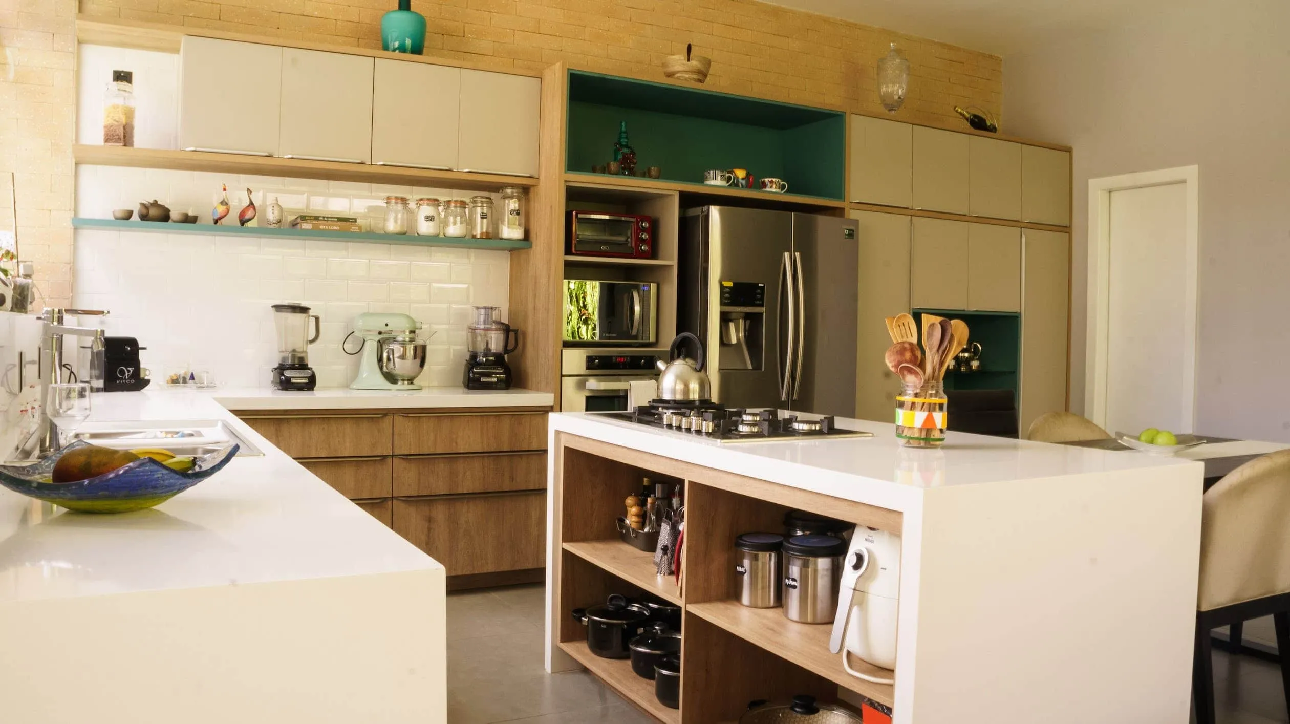 Kitchen island with an in-built pantry, a full white island which has a built-in stove on the countertop, wood laminated cabinets, seating arrangement with a table and two chairs which has a plate of sweet limes on it