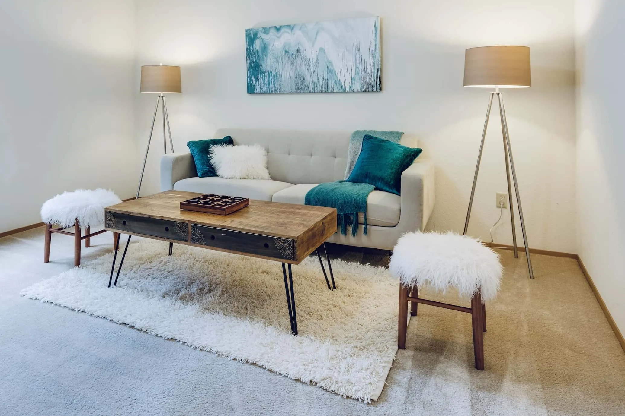reclaimed wood centrepiece, with metal legs, rugged carpet, grey sofa, white and aqua green cushions, long lamps with rustic touch, rugged cushioned stool with sleek wooden legs, white walls, painting on the wall, minimal vintage style decor