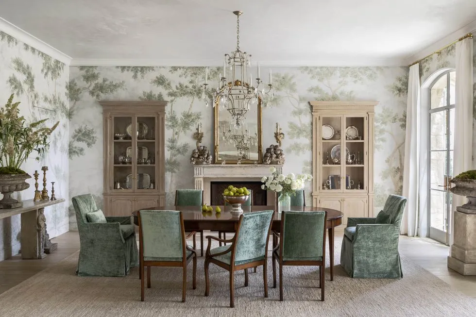 Foliage green and white dining room with a chandelier hanging over head a dining table set along with two wooden cupboards and a fire place in between them
