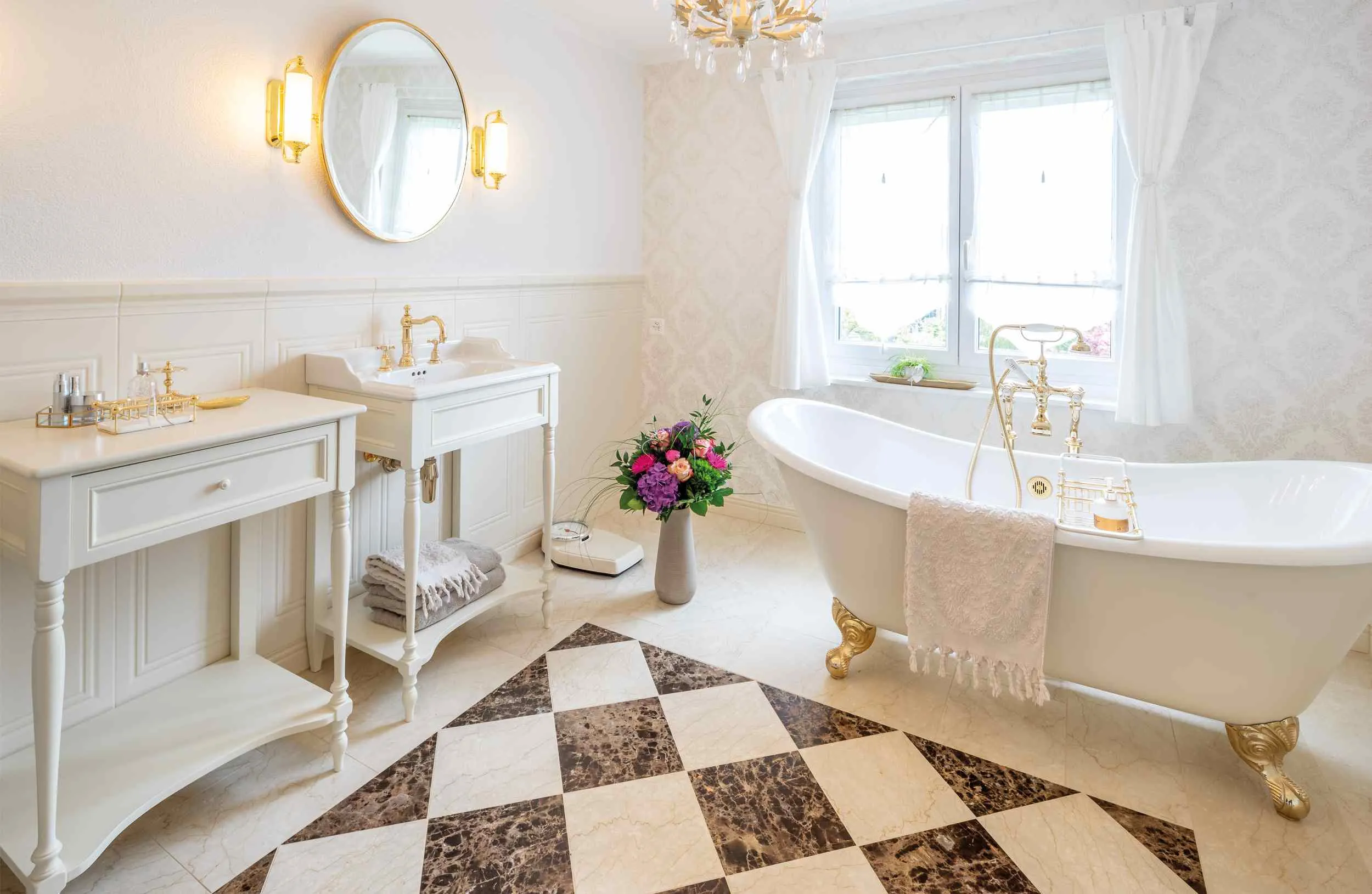 white bathroom with checkered floors, white bathtub, cabinets, table, mirror and chandelier