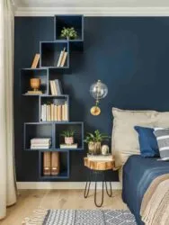 bedroom with blue accent wall, neutral adjacent walls, white ceiling, wooden floor, beige curtains, geometric wooden frames on the blue wall holding books, décor items, small plants, wooden stool next to the bed holding plants, white and blue pattern rug under the bed,